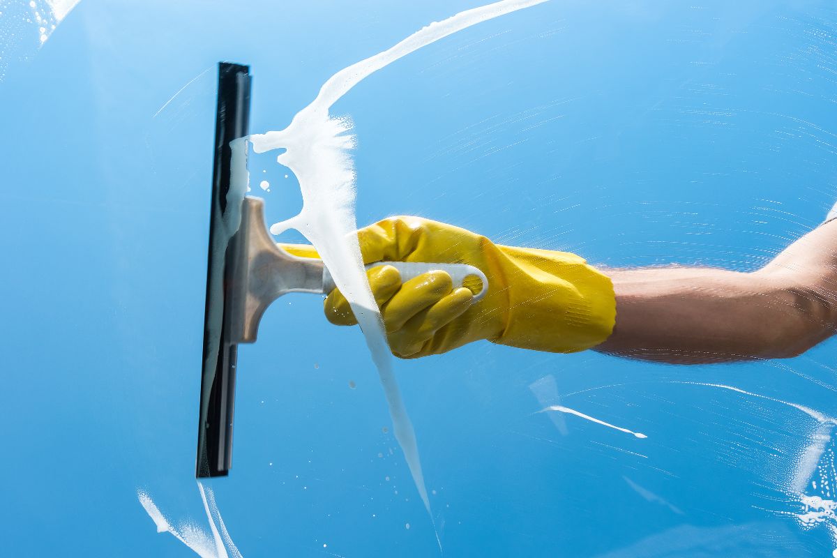 A man wearing yellow gloves cleans a car's windshield with a window cleaner, focusing on removing dirt and smudges.